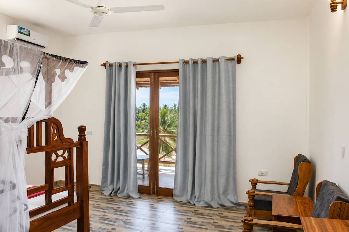 A room with a bed, a wooden chair, a table, a ceiling fan, and a view through a glass door.