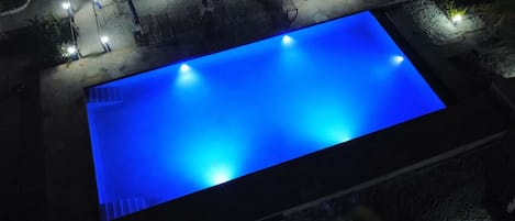 Piscina rectangular con un ligero tono azul, rodeada por un sendero con bancos e iluminación.