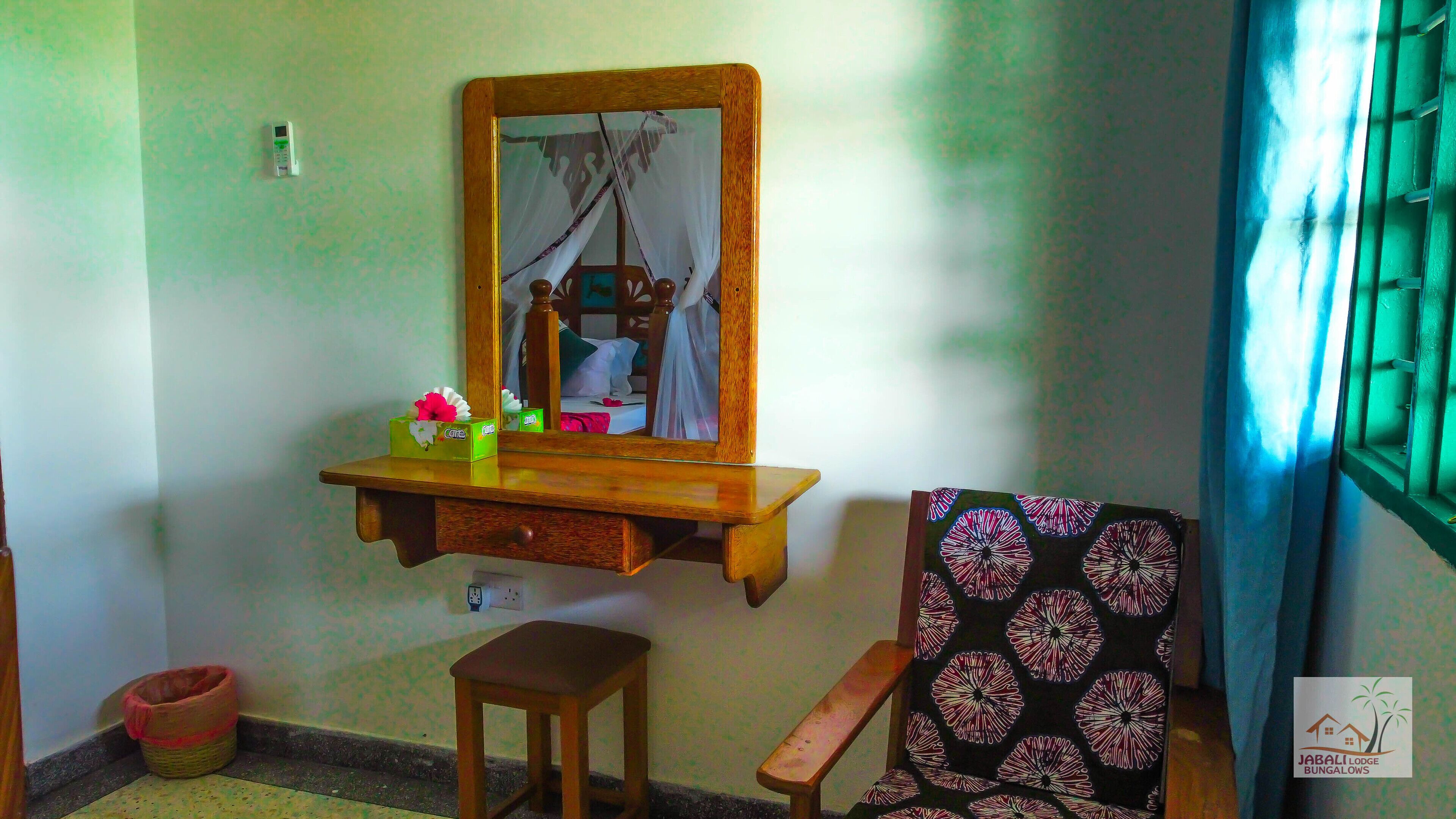 Une pièce comprenant une coiffeuse en bois, un miroir, un petit tabouret en bois, une chaise à motifs et une fenêtre avec des rideaux.
