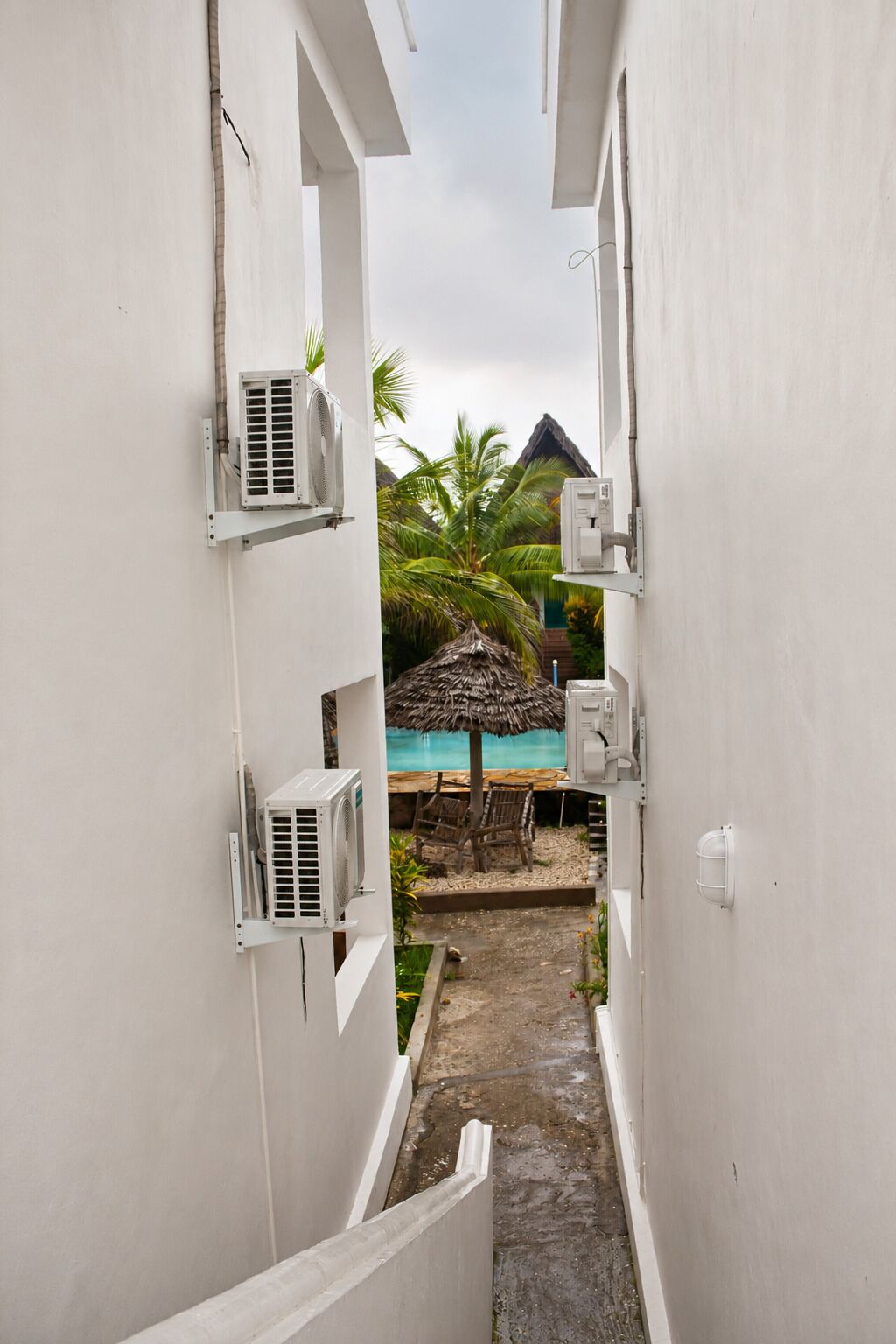 A narrow pathway leading to a pool area with a thatched umbrella and lounge chairs.