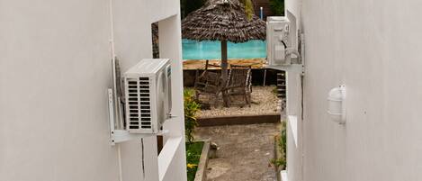 A narrow pathway leading to a pool area with a thatched umbrella and lounge chairs.