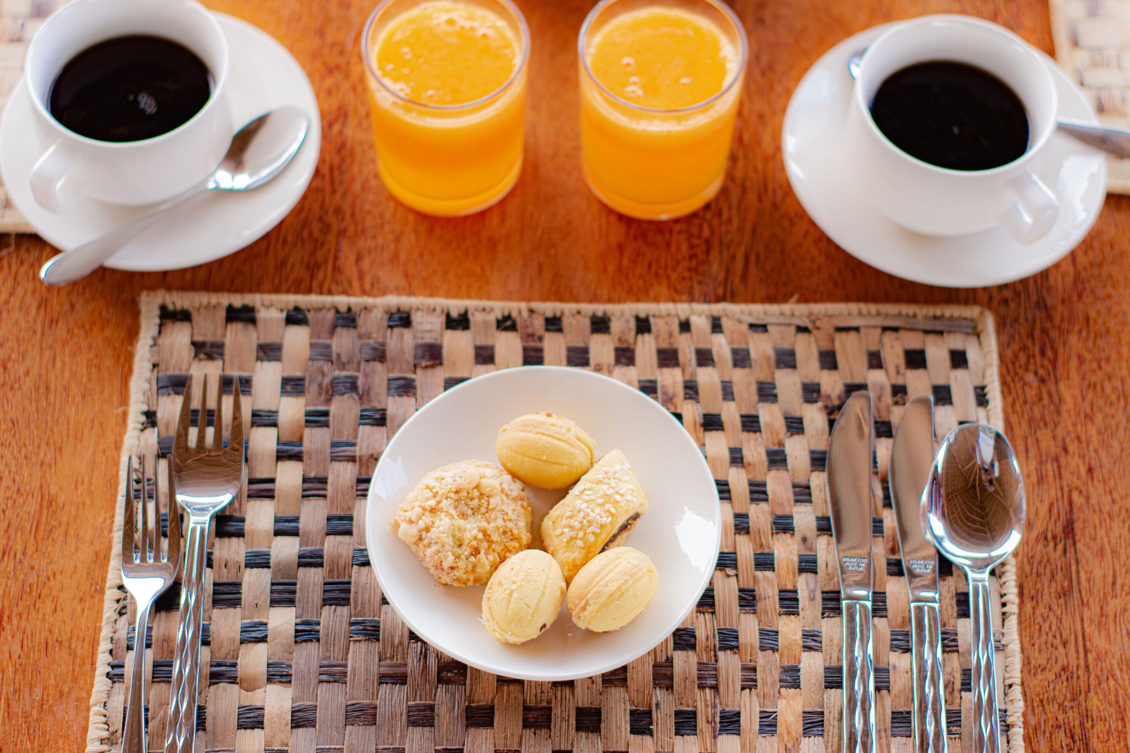 Un desayuno con un posavasos tejido, un plato de pasteles y dos tazas de café con un vaso de jugo de naranja.
