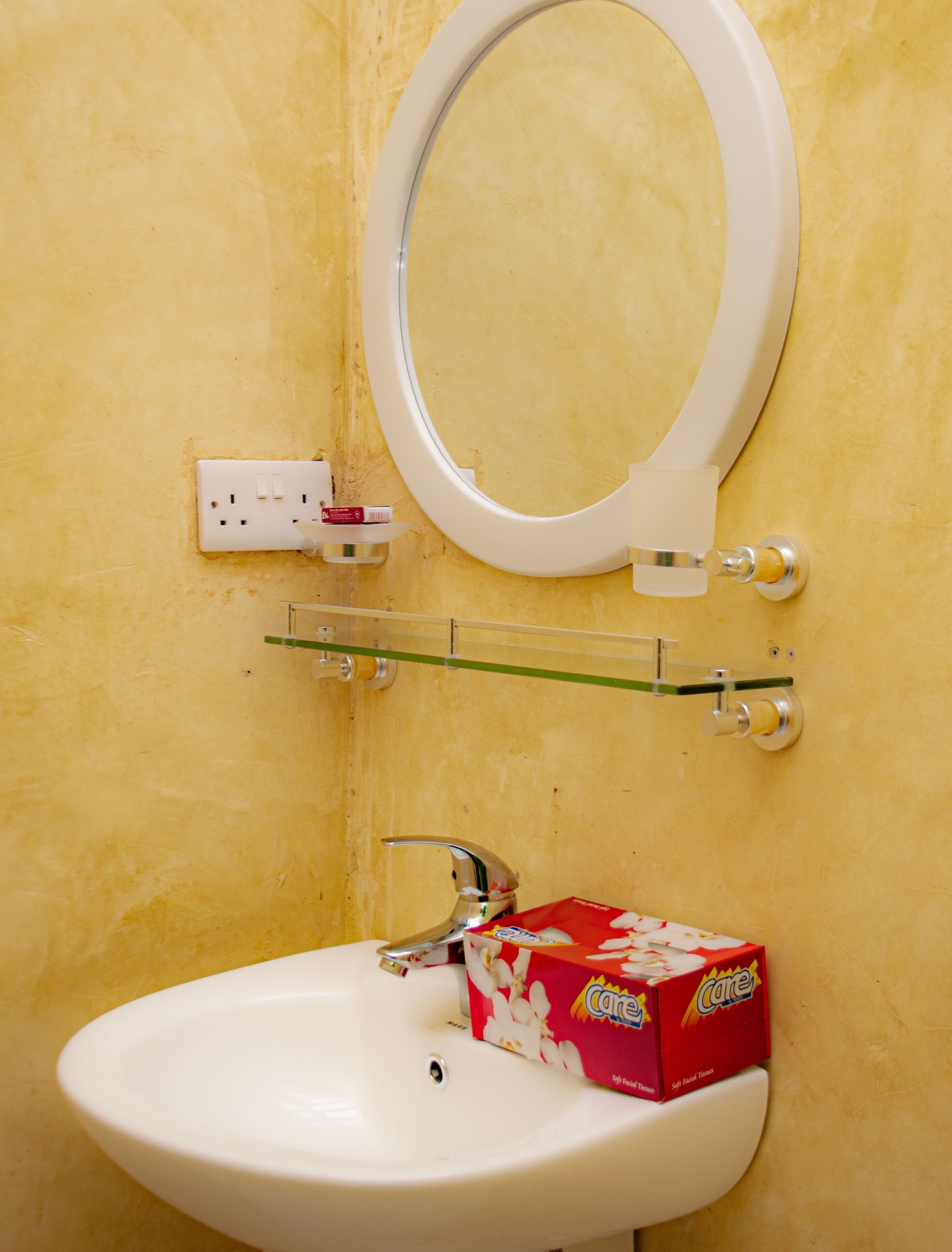 A bathroom with a white sink, a mirror, and a toilet. There is a glass shelf above the sink holding a box of tissues and a small container.