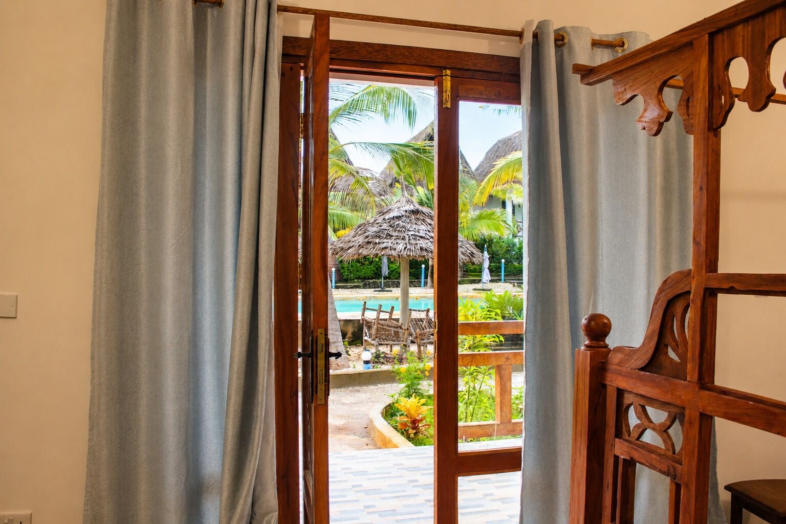 Une porte ouverte donne sur un espace piscine agrémenté d’un parasol en chaume, de meubles en bois et de plantes tropicales.