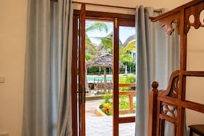 Une porte ouverte donne sur un espace piscine agrémenté d’un parasol en chaume, de meubles en bois et de plantes tropicales.