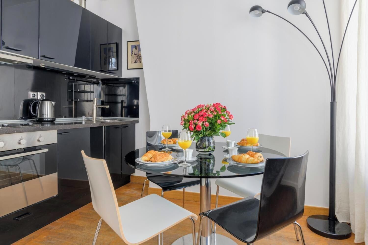 A modern kitchen with black cabinets, a stove, and a coffee maker. There is a dining table with chairs, a vase of flowers, and plates of food.
