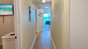 A hallway with a television displaying 'Legacy Vacations' and a painting of a beach. - Palms 204 Beachfront Condo with Free Beach Service! (Fort Walton Beach)
