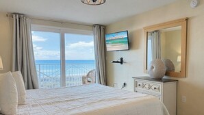 A bedroom with a large bed, a dresser, a television on the wall, and a view of the ocean through the window. - Palms 204 Beachfront Condo with Free Beach Service! (Fort Walton Beach)