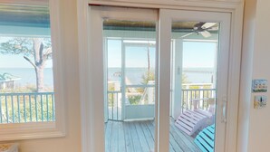 A view from a room with a large window overlooking a wooden deck with a railing, a ceiling fan, and a view of the ocean and a pier. - Bayfront with Gulf and Pool Access (Port Saint Joe)