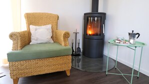 Un fauteuil en osier avec un coussin vert, une petite table avec une cafetière et un poêle à bois.