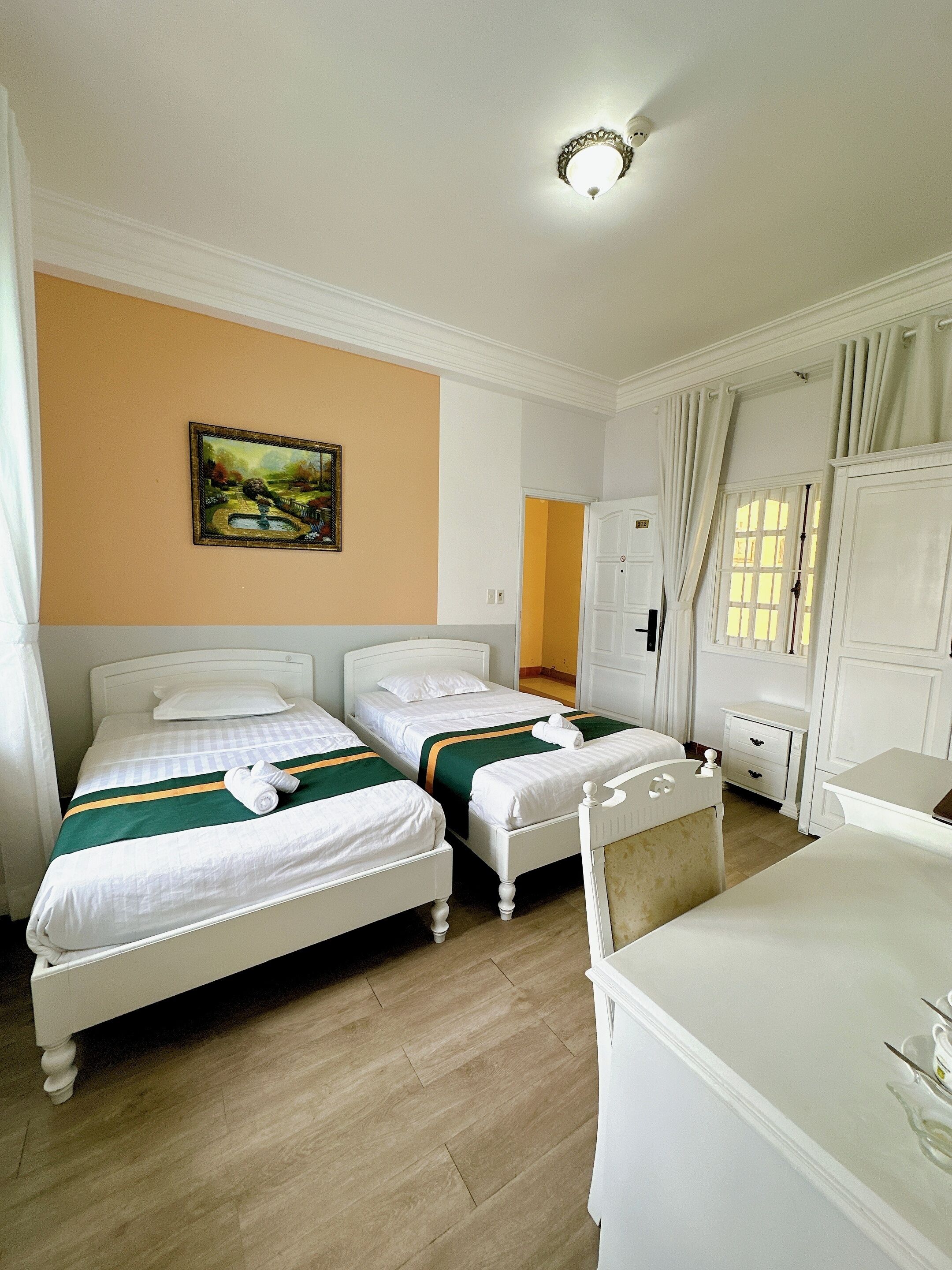 A hotel room with two single beds, a painting on the wall, a desk, and a window with curtains.