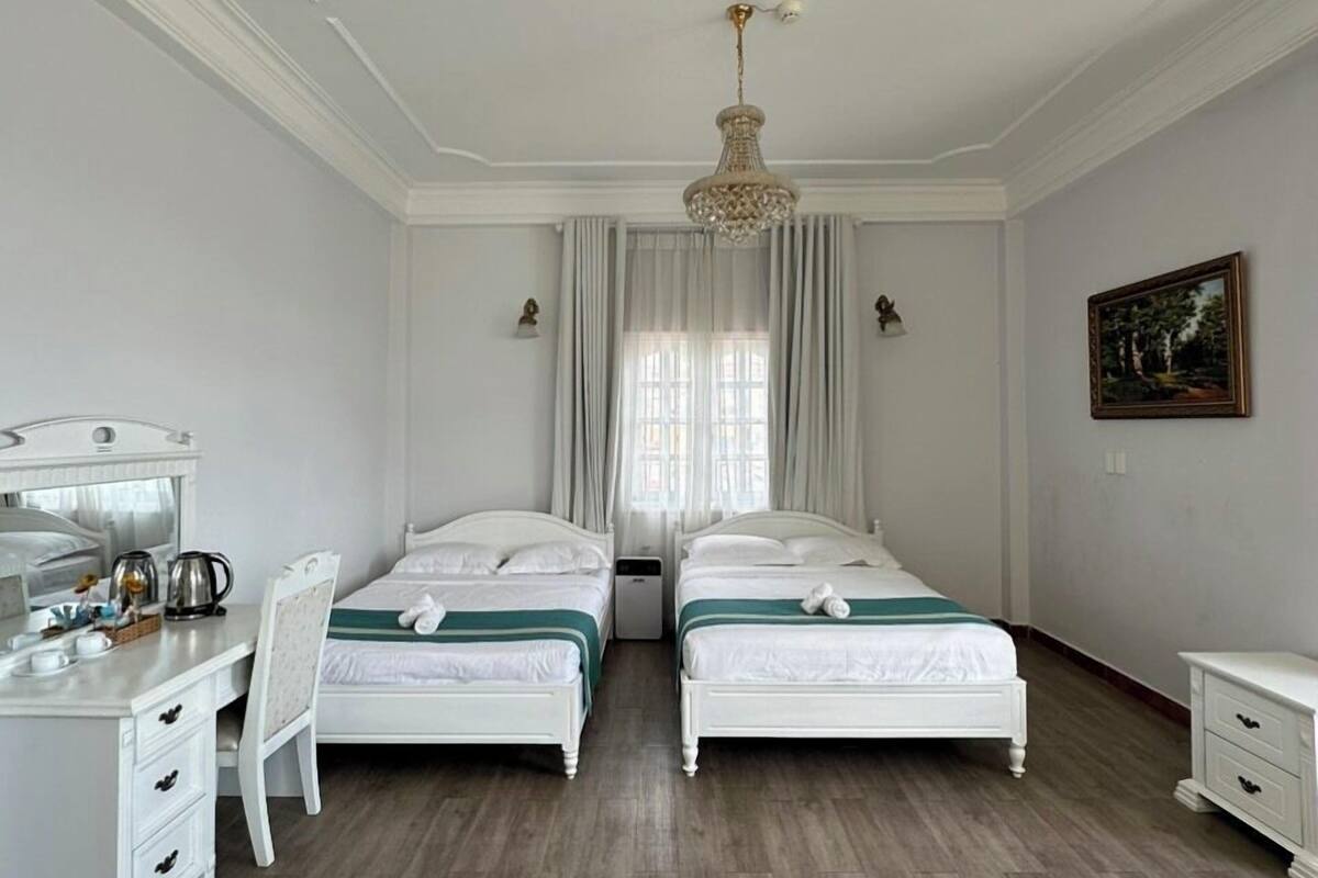 Hotellihuone, jossa on kaksi erillistä sänkyä, työpöytä tuolilla, pieni yöpöytä ja suuri ikkuna, jossa on verhot.