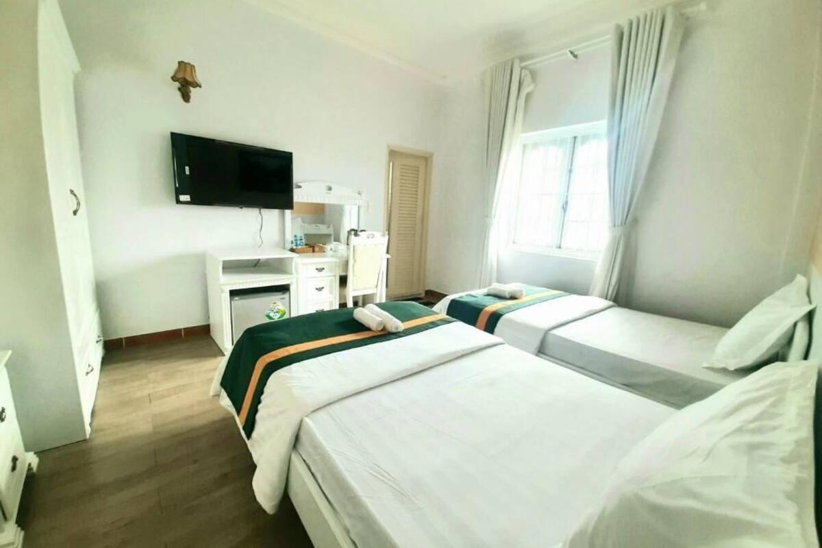 Hotellihuone, jossa on kaksi sänkyä, seinälle asennettu taulutelevisio, työpöytä tietokoneella ja ikkuna, jossa on verhot.