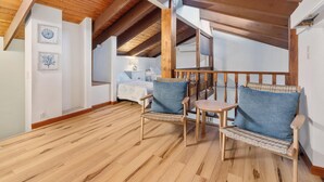A wooden-floored room with a high ceiling, a bed with a white headboard, two wicker chairs with blue cushions, and a small wooden table. - Summer Deal Kona Condo with Private Lanai Pool and Fast WiFi (Kailua Kona)