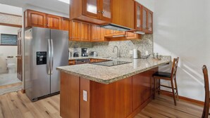 A kitchen with wooden cabinets, a stainless steel refrigerator, granite countertops, and a built-in sink. - Summer Deal Kona Condo with Private Lanai Pool and Fast WiFi (Kailua Kona)