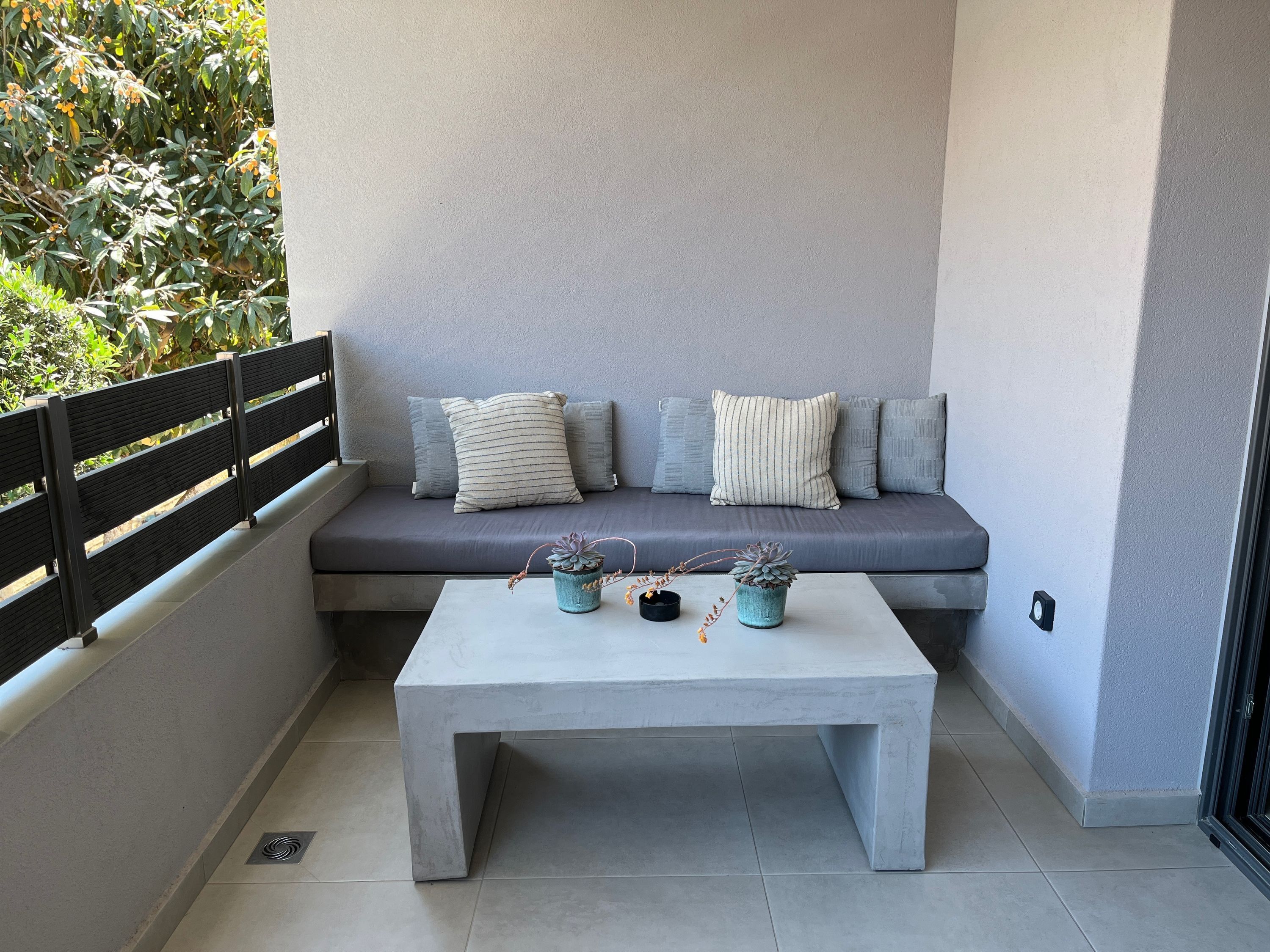 Un balcone moderno con un divano grigio, due cuscini e un tavolino in cemento con piante in vaso.