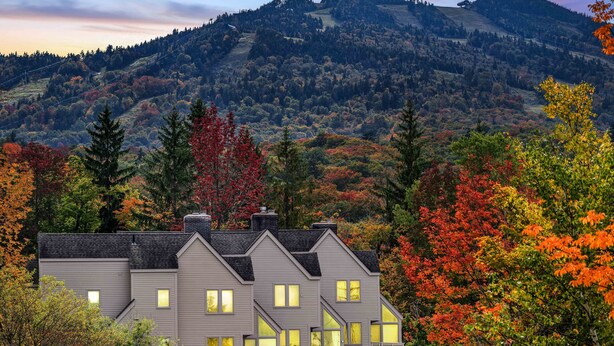 A two-story house with multiple windows and a sloped roof, surrounded by autumn-colored trees and a mountainous backdrop. - Beautiful 2br/2ba Fall Line Condo W/ Mountain Views, Pool, Hot Tub, Sauna, Shuttle. G6b 2 Bedroom Condo (Killington)