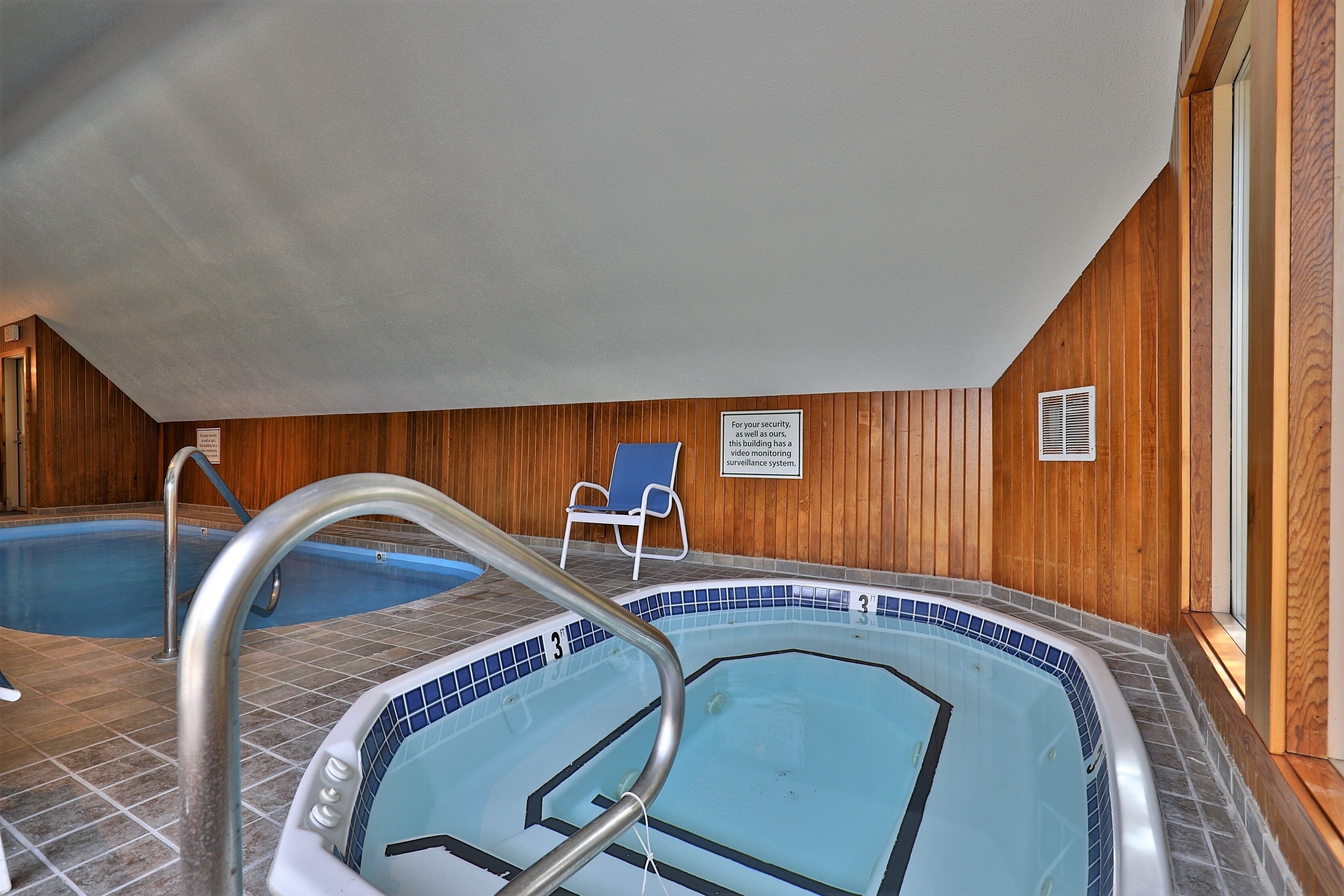 An indoor swimming pool with a blue tiled floor, a metal pool ladder, and a wooden wall with a surveillance sign.