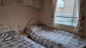 A bedroom with two beds, each covered with grey and white bedding featuring sloth patterns. There is a window with blinds and floral curtains. - Caravan - Sleeps 4 - Pet Friendly - Parking (Newton Stewart)