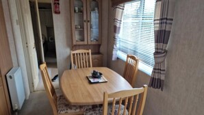 A dining area with a wooden table and four chairs, a cabinet with glass doors, and a window with blinds. - Caravan - Sleeps 4 - Pet Friendly - Parking (Newton Stewart)