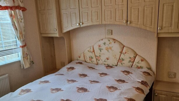 A bedroom with a bed, floral headboard, and light-colored wooden cabinets above. - Caravan - Sleeps 4 - Pet Friendly - Parking (Newton Stewart)