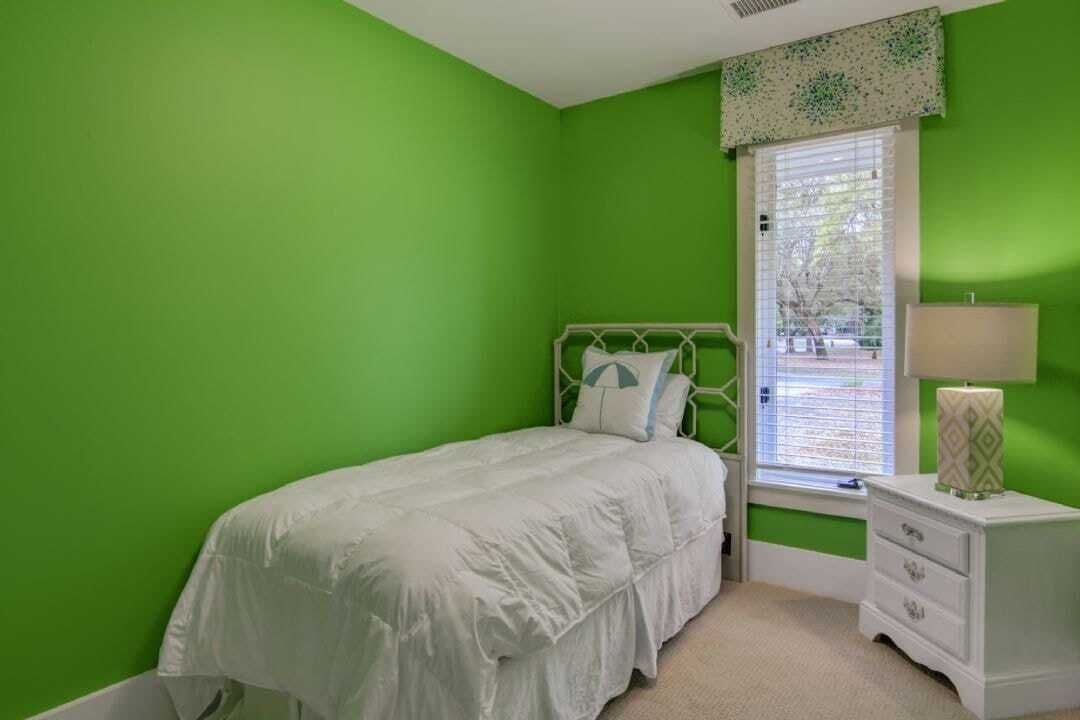 A bedroom with a green wall, a white bed, a window with blinds, and a bedside table with a lamp.
