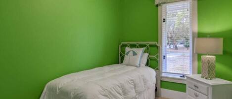 A bedroom with a green wall, a white bed, a window with blinds, and a bedside table with a lamp.