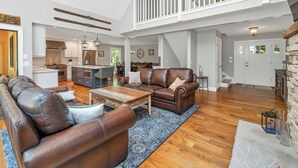 A spacious living area with a brown leather sofa, a wooden coffee table, and a dining table with chairs. A kitchen with modern appliances is visible in the background.