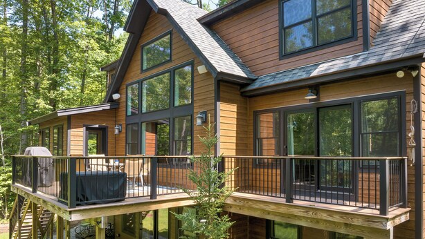 A two-story wooden house with large windows, a deck, and a covered outdoor seating area.