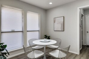 A dining area with a round table, four wireframe chairs, and two large windows with blinds.