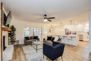 A modern living room with a sofa, coffee table, and a TV. There is a kitchen area with a bar and white cabinets.