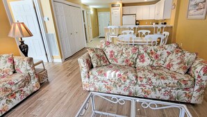 A living room with a floral sofa, a dining table with chairs, and a kitchen area in the background.