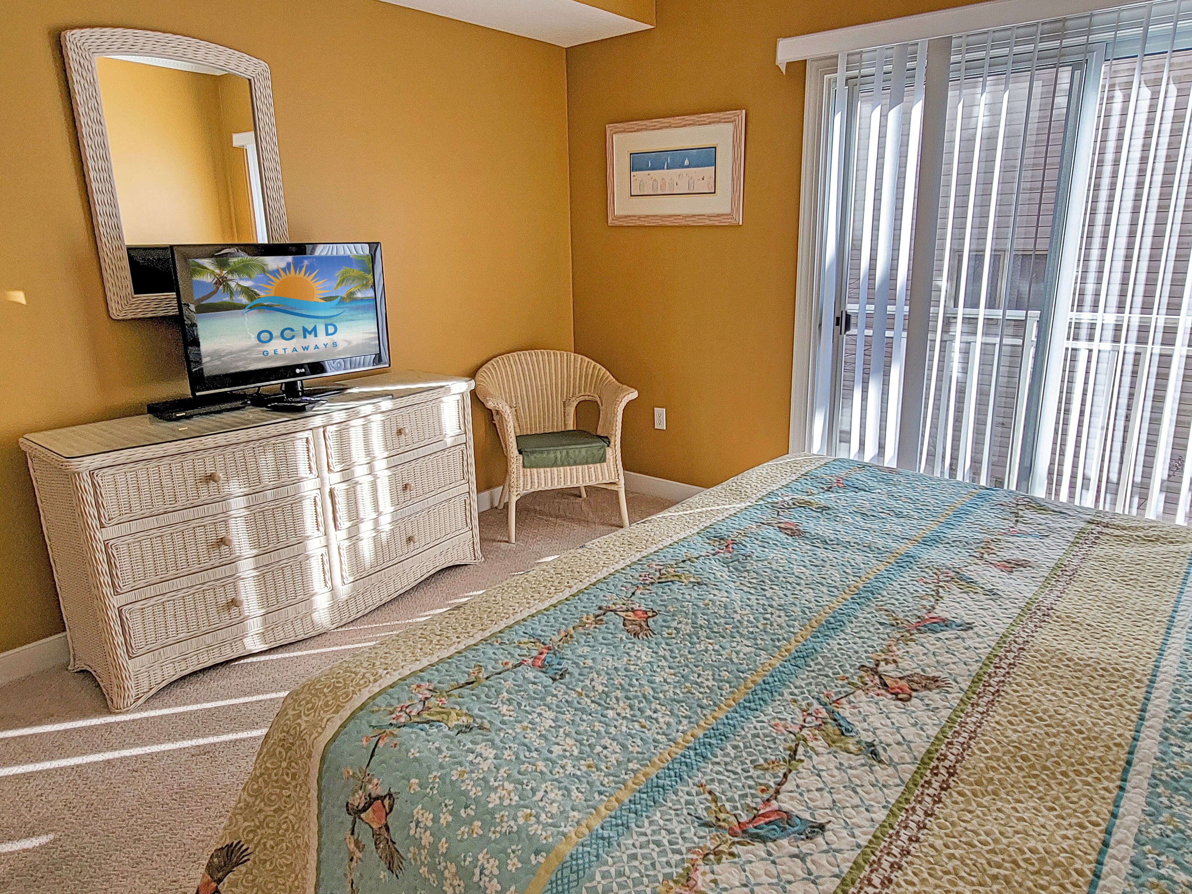 Une chambre avec un grand lit, un téléviseur posé sur une commode, un fauteuil en osier et une fenêtre avec des stores.