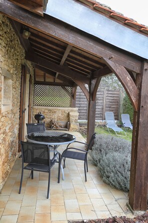Een overdekt terras met een ronde tafel en stoelen, een barbecue en een stenen muur.