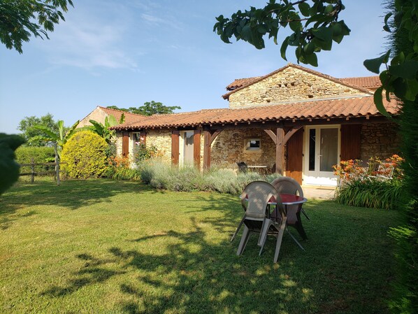A stone house with a tiled roof, a patio with chairs, and a well-maintained garden.