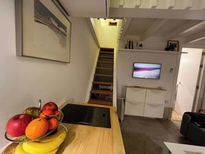 Un salon moderne avec une table en bois sur laquelle sont disposés des fruits, un téléviseur à écran plat fixé au mur et un escalier menant à un autre niveau.