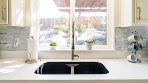 A kitchen with a modern faucet, a window with a view of a backyard, and a countertop with a paper towel holder and a mug rack. - Bluebird Day - Mountain Escape, Cozy a 3-bedroom home in Bend, OR. (Bend)