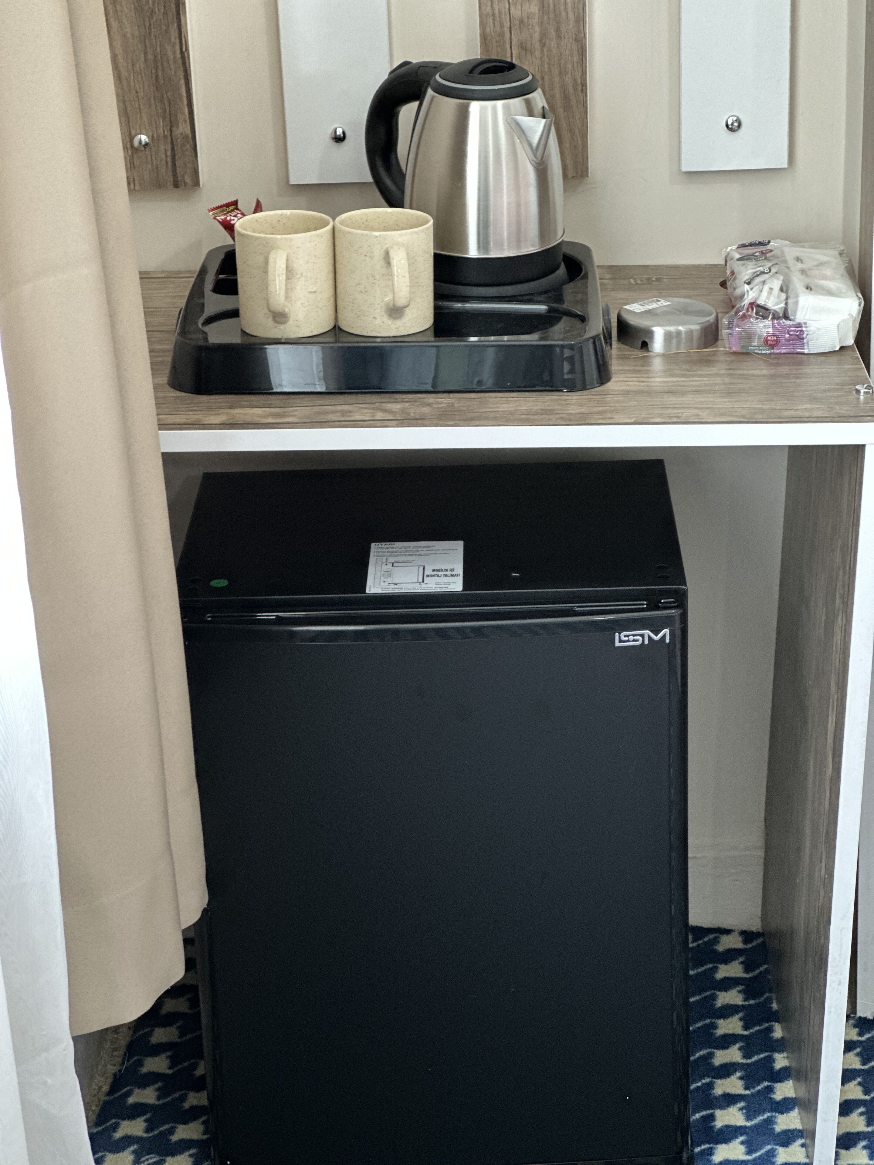 A kitchenette with a black refrigerator, a kettle, and two mugs on a countertop.