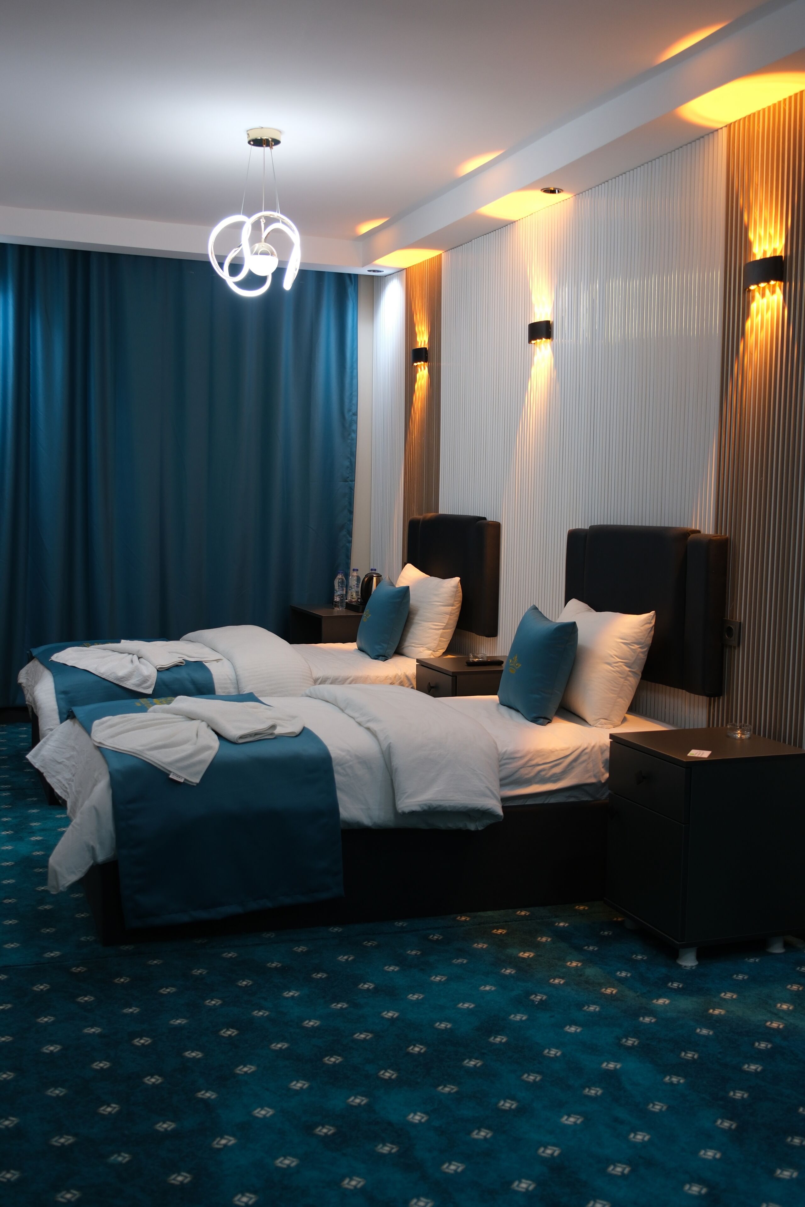 Habitación de hotel con dos camas, un ventanal con cortinas, una lámpara de araña moderna y lámparas de pared.