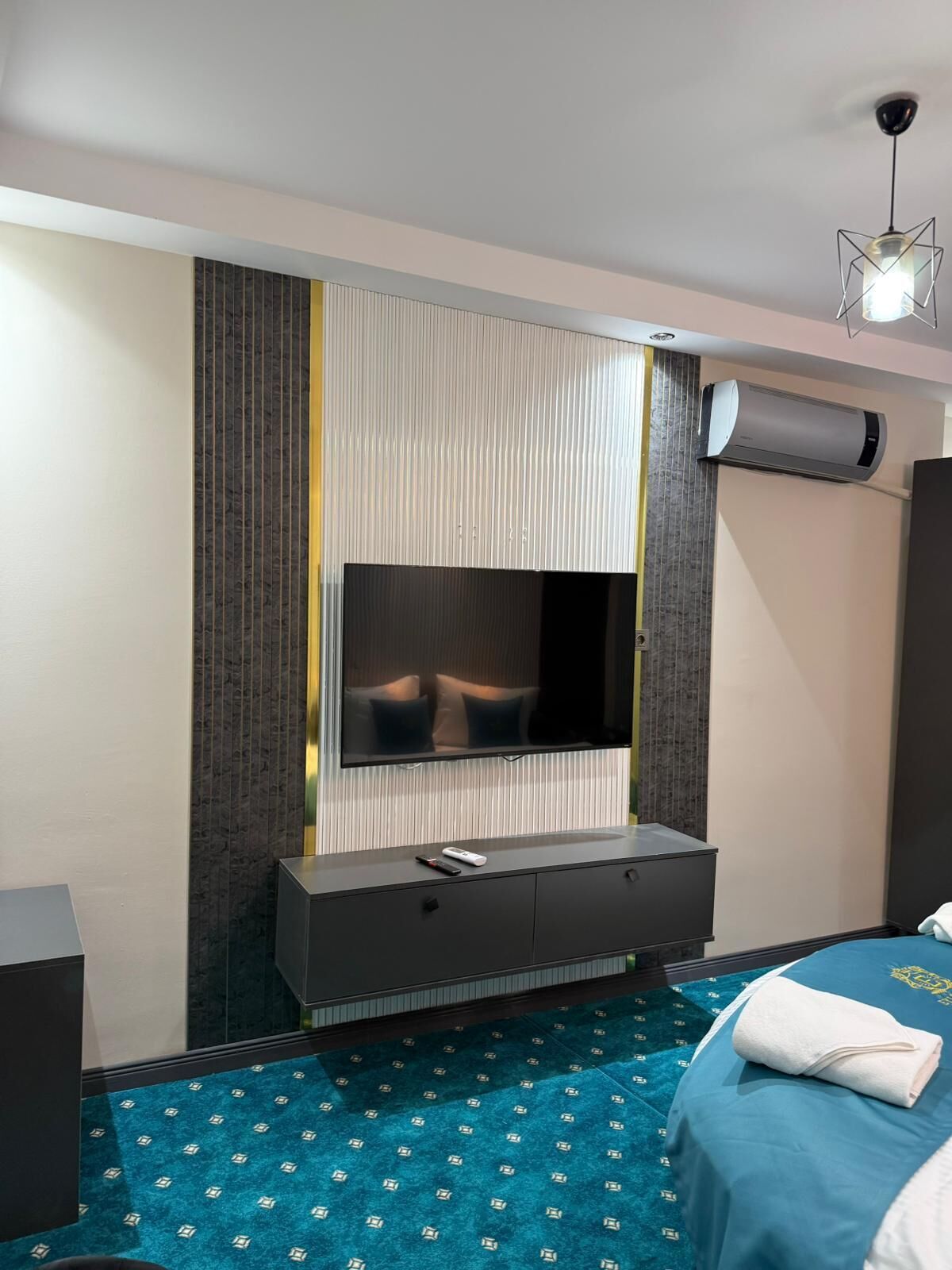 A modern hotel room with a large flat-screen TV mounted on a textured wall, a dark-colored console table with a remote control, a blue patterned carpet, and a bed with a blue bedspread and a white towel.