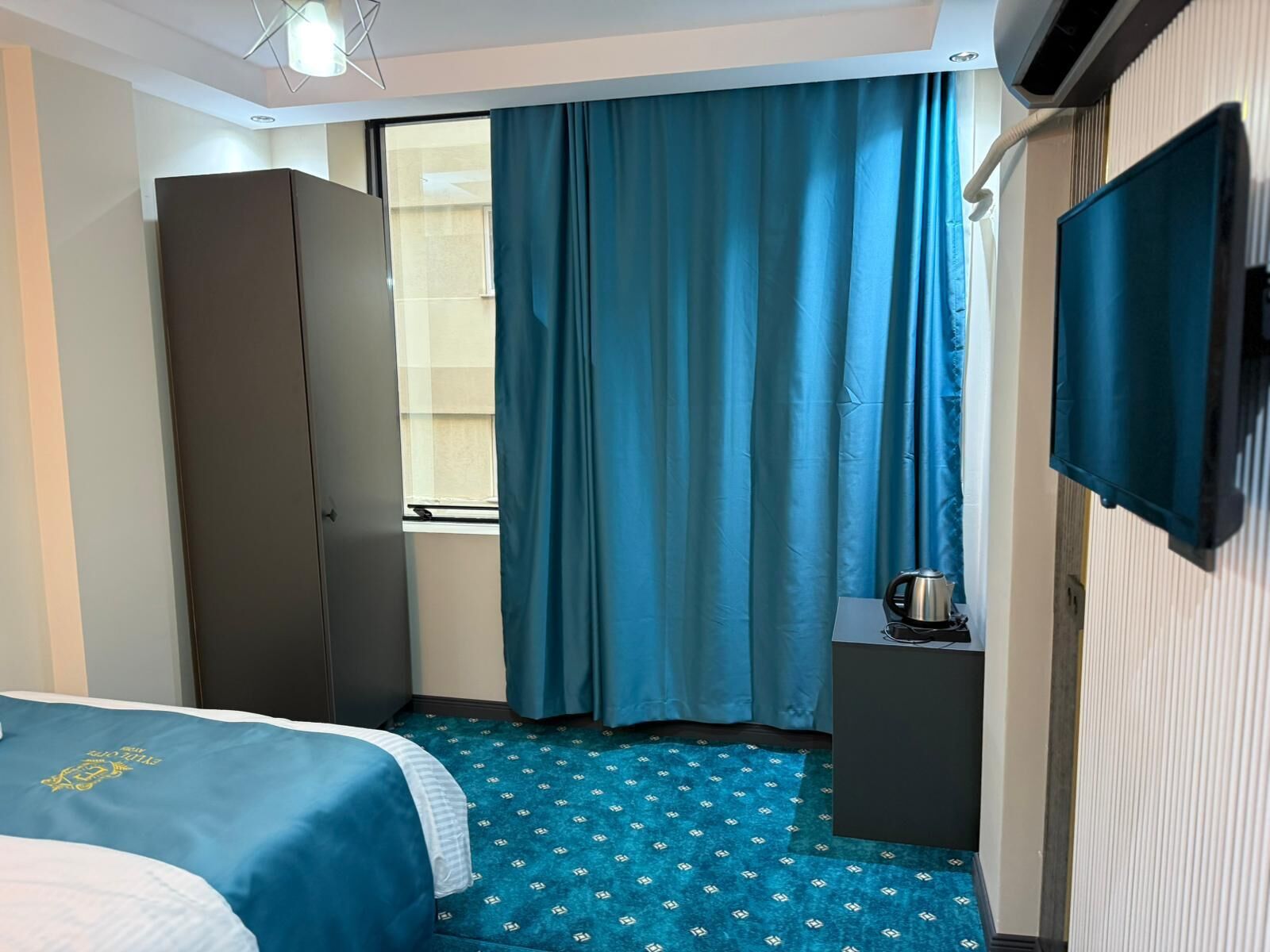 A hotel room with a bed, a wardrobe, a small table with a kettle, and a television.