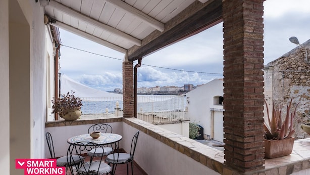 A balcony with a table and chairs, a potted plant, and a view of the sea and buildings.