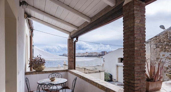 A balcony with a table and chairs, a potted plant, and a view of the sea and buildings.
