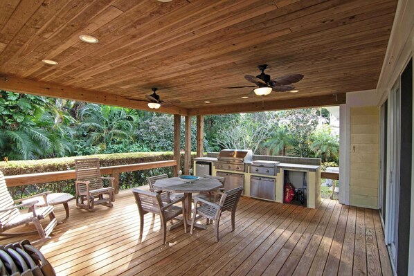 An outdoor wooden deck with a table and chairs, ceiling fans, and an attached kitchen area.