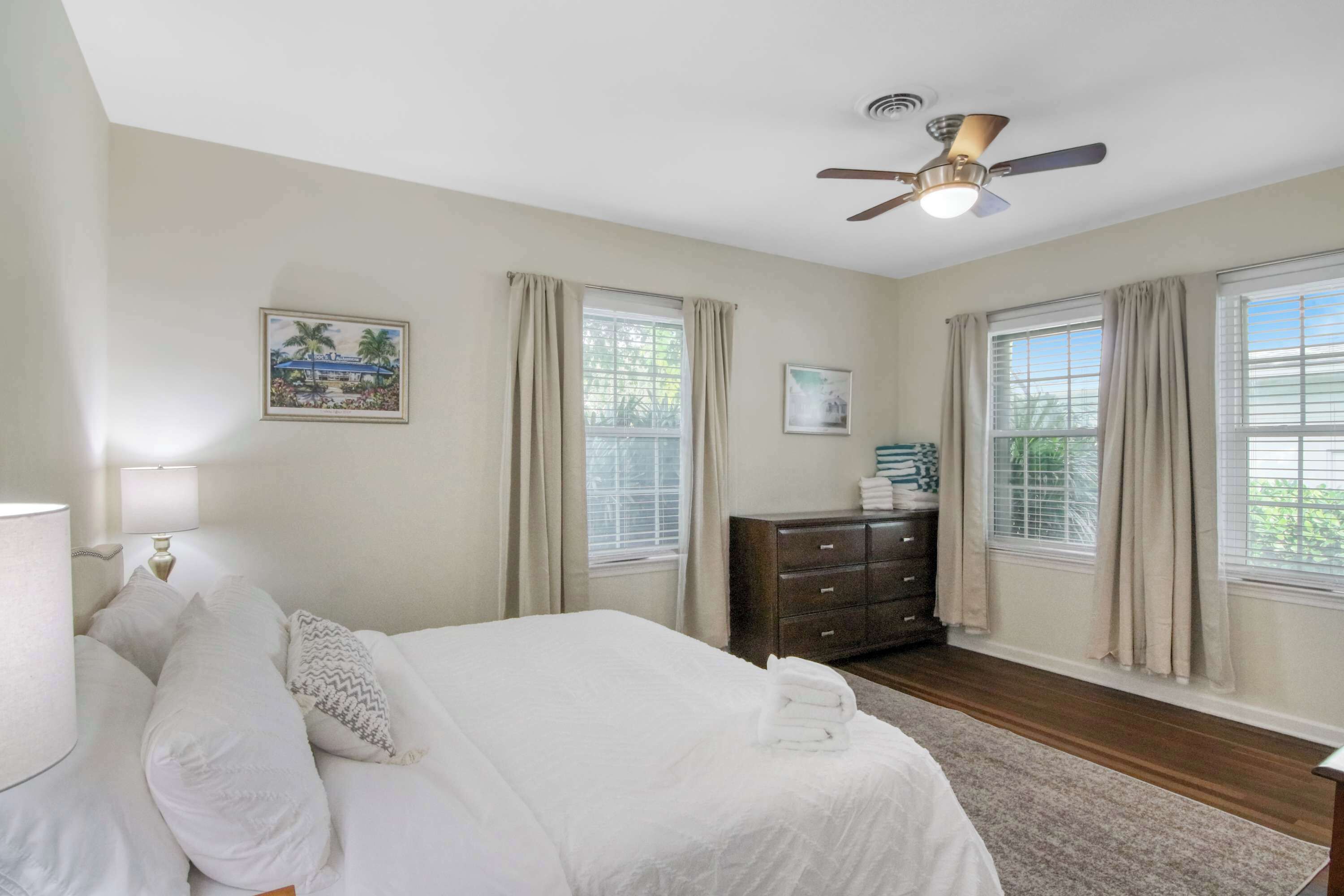 A bedroom with a bed, a dresser, a ceiling fan, and two windows with curtains.