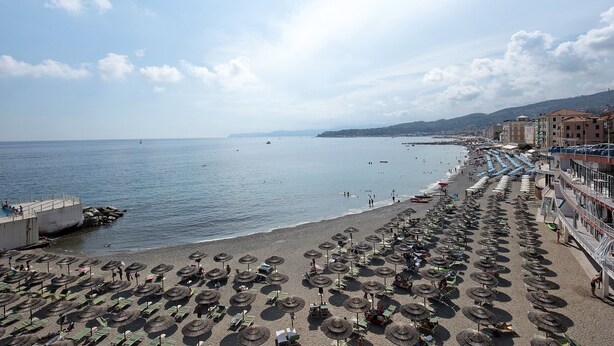 Une plage bordée de rangées de parasols et de chaises longues, une jetée et des bâtiments le long de la côte.
