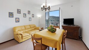 Un salon avec un canapé jaune, une table à manger recouverte d’une nappe jaune, un lustre, un téléviseur posé sur un meuble en bois et des tableaux encadrés accrochés au mur.