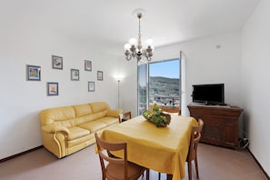 Ein Wohnzimmer mit einem gelben Sofa, einem Esstisch mit gelber Tischdecke, einer Kristallleuchtre, einem Fernseher auf einer Holzkommode und gerahmten Bildern an der Wand.