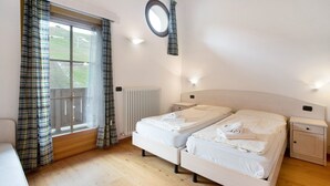 A bedroom with two beds, a window with curtains, a bedside table, a nightstand, and a radiator. - Apartment 'Baita Valandrea 3' with Mountain View, Shared Garden and Wi-Fi (Livigno)