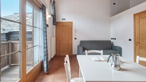 A room with a wooden-framed window, a white table with a metal coffee pot, a grey sofa, and a wooden door. - Apartment 'Baita Valandrea 3' with Mountain View, Shared Garden and Wi-Fi (Livigno)